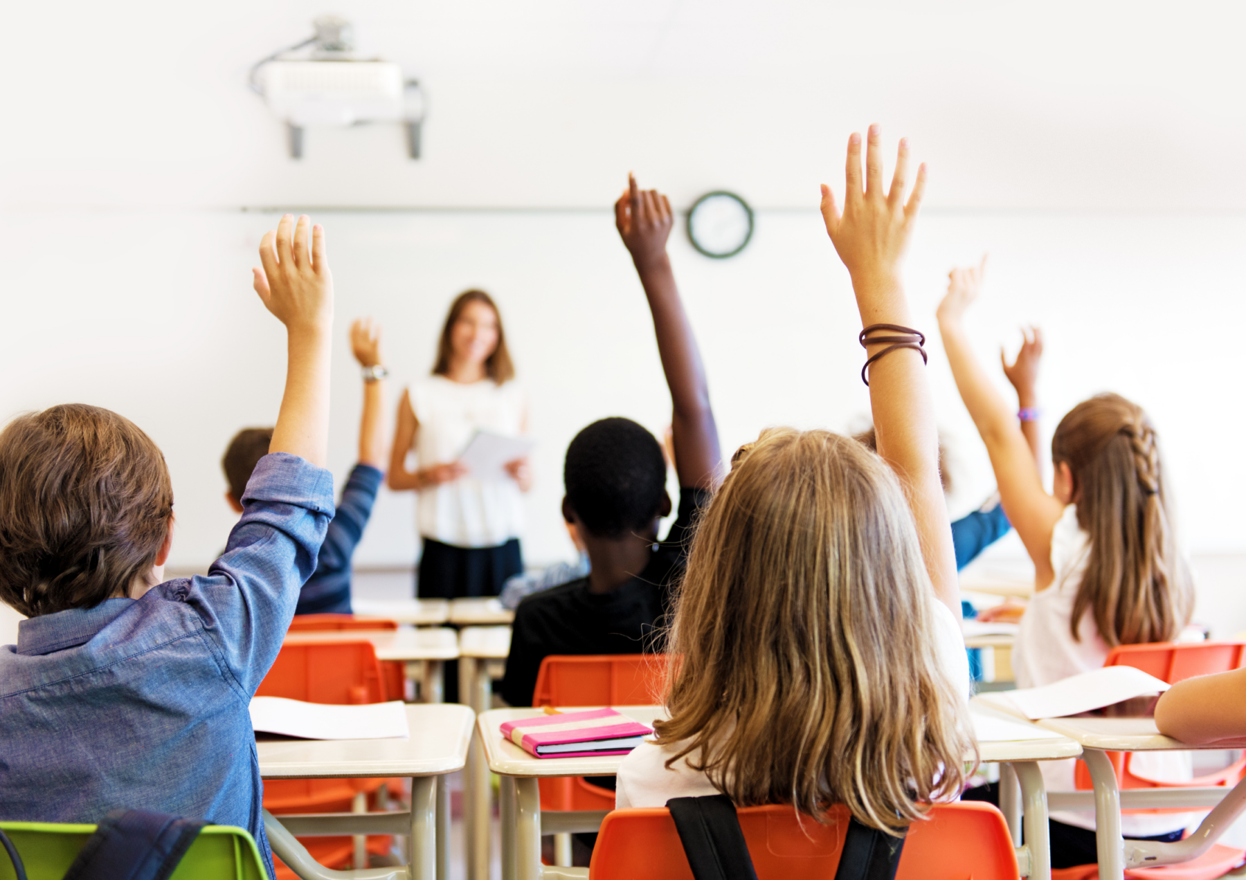 school classroom with students raising their hands with teacher at the front of the class. 